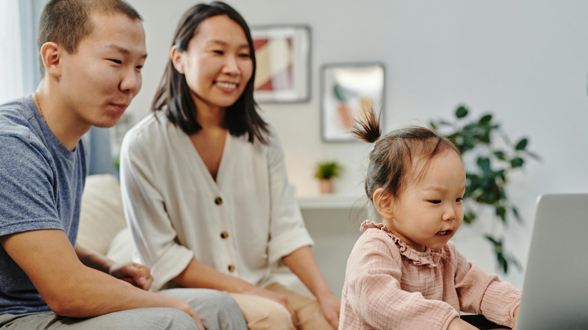 A father and mother sit at home with their young daughter as she engages with a laptop