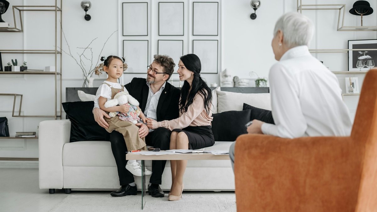 A young girl holding a stuffed bunny sits with her parents on a couch during a therapy consultation in a warm home setting