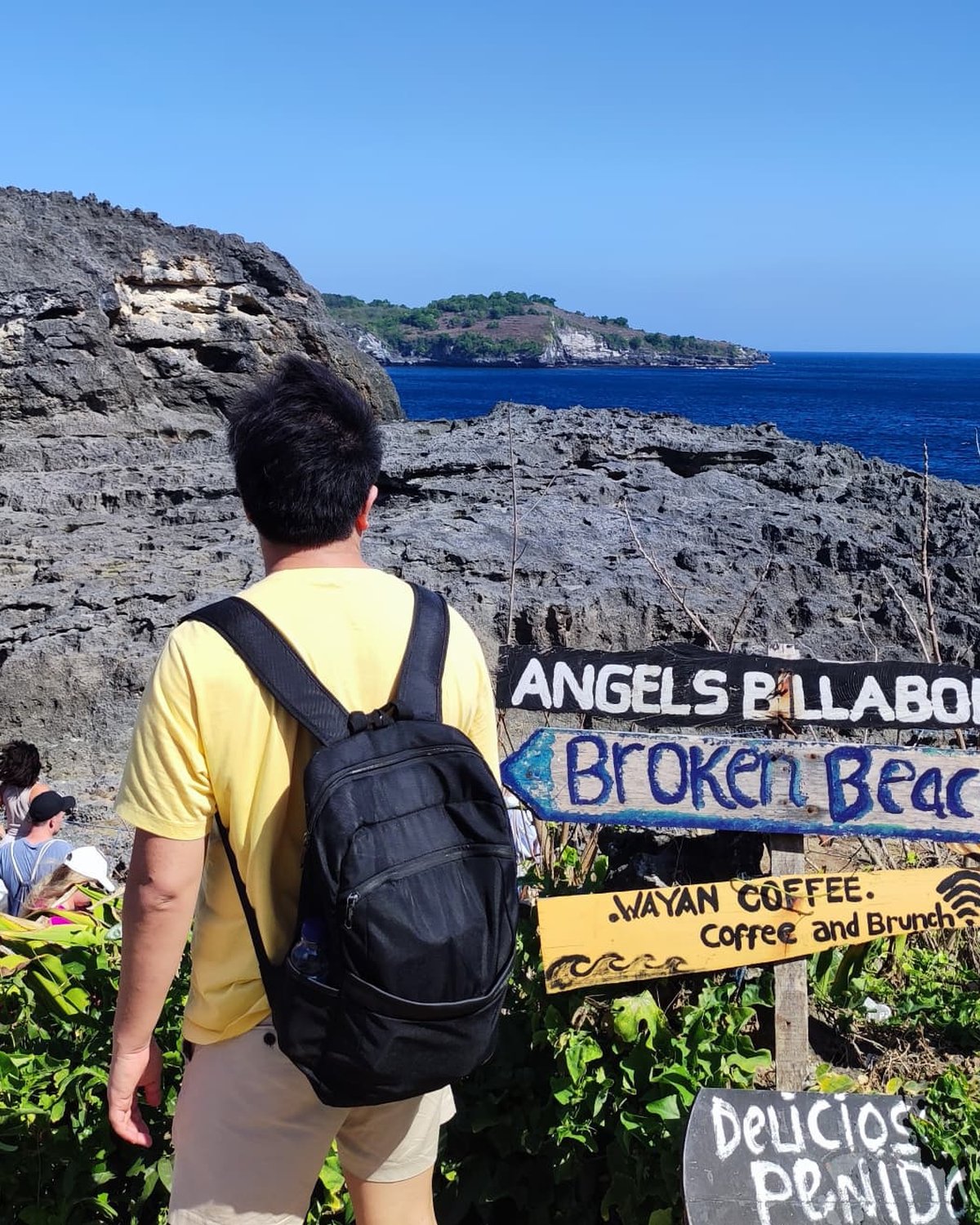 The founder of Curauti, photographed from behind on a clifftop trail in Nusa Penida, Indonesia, looking out toward the ocean and a small island in the distance, with hand-painted Angels Billabong, Broken Beach, and Wayan Coffee signs nearby.